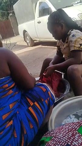 Two women are washing clothes outside. One woman wears a blue and orange patterned dress, while the other wears a light-colored t-shirt and dark shorts. Both women sit on low stools, with metal basins of water and clothes in front of them. They scrub and wash the clothes by hand. A white pickup truck is parked in the background. The ground appears to be dirt or gravel.