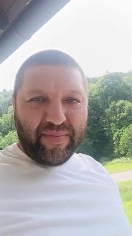 A man with short brown hair and a beard, wearing a white t-shirt, smiles at the camera. He is outdoors, with a background of green trees and a bright sky. A trampoline can be seen in the background.