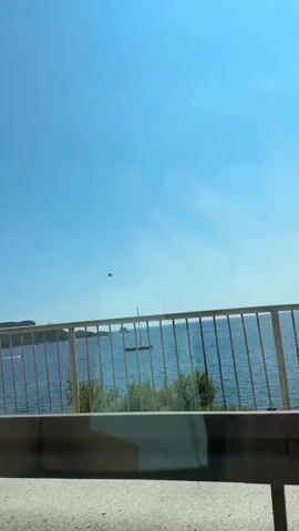 The video displays a clear blue sky above a body of water, likely the sea. In the foreground, a white railing runs horizontally across the bottom of the frame. Beyond the railing, a green tree with dense foliage is visible on the left side. The water is calm with several boats scattered across it. A sailboat with a mast is prominent in the middle ground. In the sky, a person is parasailing, attached to a red parachute, with a boat visible towing them. The overall scene suggests a bright, sunny day at a coastal location, likely a resort or a tourist destination.