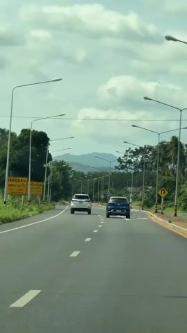 The video shows a POV shot from a vehicle driving on a paved road with multiple lanes.  Two cars, a white SUV and a blue sedan, are traveling ahead in the same direction. A motorcycle carrying two people is also visible on the right side of the road. The road is lined with streetlights and trees. In the background, hills or mountains are visible under a cloudy sky. Road signs are present along the side of the road, including one with a yellow background and text in a foreign language, and another indicating a U-turn. The road has white lane markings and a yellow border.