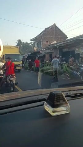 The video shows a street scene in what appears to be a market area. The foreground shows the interior of a vehicle, with a dashboard ornament visible. The street is busy with traffic and pedestrians. There are several vehicles, including a yellow truck, a white tanker truck with "PERTAMINA" written on it, and several motorcycles. People are seen interacting with each other and with goods that appear to be produce, such as bananas and coconuts, being loaded onto or unloaded from motorcycles and carts. Buildings line the street, some of which appear to be shops or market stalls. The overall atmosphere suggests a bustling marketplace with commercial activity.