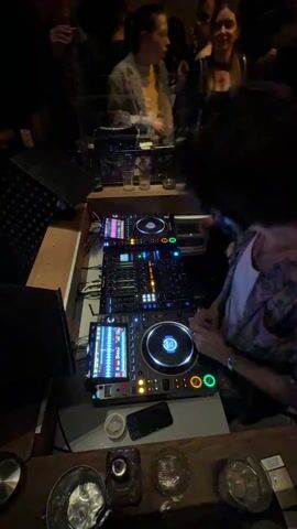 The video shows a DJ working behind a deck at a party or club. The DJ is operating two Pioneer CDJ-3000 media players and a Pioneer DJM-A9 mixer, all arranged horizontally on a wooden countertop. Several drinks and a phone are beside the equipment. The DJ is wearing a patterned shirt and a watch on their left wrist. Their hair is black. The video is shot from a high angle, looking down at the DJ and the equipment. People can be seen standing and chatting in the background. The lighting is dim, with the equipment illuminated by its own lights.