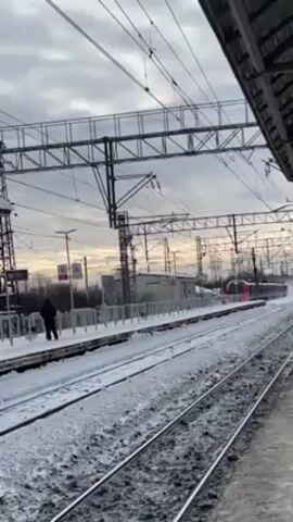 The video shows a train arriving at a snowy train station on a cloudy day. The train is red and gray. There is a person standing on the platform. The camera is positioned under a covered area. Wires and metal beams are visible overhead.