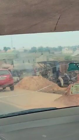 The video captures a dynamic street scene in Uke, Nigeria, viewed from inside a moving vehicle through a cracked and somewhat dusty windshield. The time of day appears to be daylight, with an overcast or hazy sky suggesting either a cloudy day or a dusty environment.

The scene is bustling with activity, featuring numerous people and vehicles. Several yellow tricycles, commonly known as Keke Napeps, are prominent, seen moving, parked, or with people getting in and out. Other cars are also visible, including a red car at the beginning of the video that briefly stops with its door open. People are seen walking on the roadside, some crossing the street, and others appearing to be waiting for transport or engaged in daily activities.

The setting includes buildings in the background, characteristic of an urban or semi-urban area. The road is paved, but the shoulders appear unpaved and dusty. In the foreground, inside the filming vehicle, a blue plastic bucket is partially visible on the dashboard or hanging. Text on the bucket reads "DELTA" and some smaller, unreadable lines below it. The ambient sound includes general street noise, engine sounds, and voices, indicating a lively atmosphere.