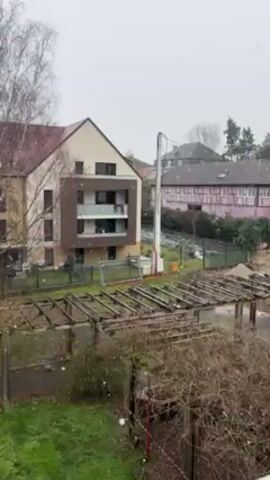 The camera pans across a light dusting of snow that has fallen on an urban scene in Germany. The camera begins by focusing on a tan, three-story building with a brown accent. Next to the building, a yard is partially covered by a brown trellis. A few green patches of grass peak out from under the fallen snow. Behind the yard, a white telescopic boom with a red base rises into the air. As the camera slowly pans to the right, other buildings come into view, including a white building with a large balcony. A lone white car is visible on the street as the camera finishes its pan. Bare trees dot the landscape. The sky is gray and overcast.