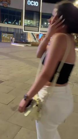 A woman in a black crop top and white pants is walking and then running away from the camera on a paved street at night. She is carrying a cream-colored purse over her right shoulder and a phone in her left hand. She turns her head to look back at the camera as she runs. In the background, there are buildings with lights, streetlights, trees, and other people.