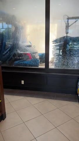 The video shows a car wash from inside a building. Large brushes are seen spinning and spraying water, cleaning a car. Cases of bottled water are stacked to the right of the car wash window. The floor is tiled.