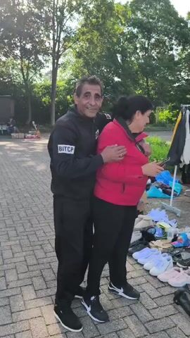 A man in a black tracksuit with "BITCP" on the sleeve embraces a woman in a red jacket. They are standing on a paved area, possibly a driveway or walkway, with trees and foliage in the background. To the right, there are various items on the ground, including shoes and clothing on racks, suggesting an outdoor market or yard sale. The man is smiling, and the woman is looking down at a phone she is holding.