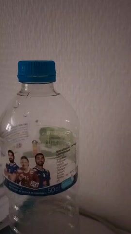 The media file shows a close-up of a plastic water bottle with a blue cap. The bottle is partially filled with water and has a label that features three male handball players in action. The label also contains text in French detailing the mineral content of the water, including Sodium, Potassium, Calcium, Magnesium, Bicarbonates, Chlorures, and Sulfates. The pH of the water is also indicated as 7.8. Below the mineral information, the text "EAU OFFICIELLE des Équipes de France de Handball" is visible, identifying the water as the official water of the French Handball Teams. A measurement of "50cl" is also present on the label.

The background of the image is a textured white wall, suggesting an indoor setting. The lighting appears to be artificial, possibly from a lamp or overhead light, casting subtle shadows. The overall mood is neutral, with no specific emotional cues conveyed. The image does not provide any clear indications of the time of day, weather conditions, or specific location beyond what is stated on the label. The focus is primarily on the water bottle and its branding.