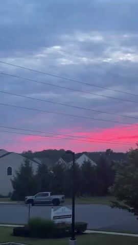 A vertical video displays a view of a suburban neighborhood at dusk. The sky is filled with clouds, some of which are tinged with bright pink and red hues, suggesting a sunset. Power lines are visible across the top of the frame. Below the sky, several houses with peaked roofs and windows are nestled among trees. In the foreground, there is a street with a sidewalk and a patch of grass. A silver pickup truck is parked on the street. A black lamppost stands near the sidewalk.