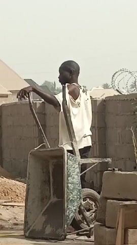 A man stands by a wheelbarrow on a construction site. He is wearing a torn white shirt and pants. He is facing away from the camera. The wheelbarrow is dirty and has a patterned cloth inside. Behind the man is a wall topped with barbed wire. A pile of sand and some cinder blocks are also visible. The sky is hazy. The sound of hammering can be heard.