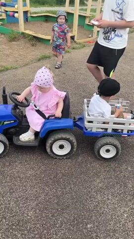 A group of children are playing on a playground. A young girl in a pink dress and hat is sitting in a blue toy tractor, holding the steering wheel. A young boy in a white shirt and black hat is sitting in a trailer attached to the tractor. An older child in a white t-shirt and black shorts is standing behind the tractor, appearing to push it. Another young child in a colorful outfit and hat is standing nearby. The playground has swings, a slide, and other play equipment. The ground is covered in asphalt and grass.
