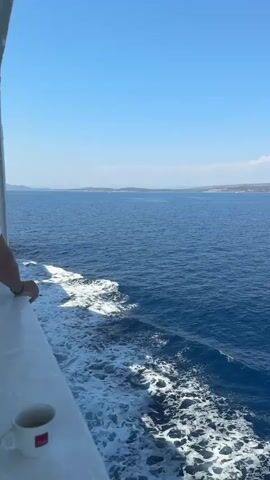 A first-person perspective video shows a person standing on the deck of a boat, looking out at the vast blue ocean. The boat is moving, creating white foam and waves in its wake. In the foreground, a white mug sits on the deck. The sky is clear and blue, and a distant shoreline with hills can be seen on the horizon. The overall atmosphere is one of calm and travel.