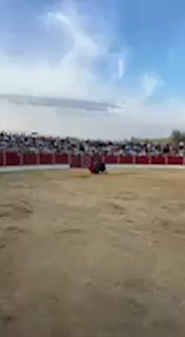 The video captures an outdoor bullfighting event in a sandy, circular arena, likely in San José del Valle, Spain.

In the center of the arena, a matador, dressed in a dark suit, stands and walks deliberately. He holds a bright red muleta (cape), making movements to engage a dark-colored bull. The bull appears calm, standing relatively still and observing the matador.

The arena is surrounded by tiered red barriers and stands, which are packed with a large crowd of spectators. Many people are visible in the stands, some wearing white or light-colored clothing, suggesting a warm, daytime event. The sky above is bright blue with scattered white clouds.

Audible cheers and applause from the crowd create an enthusiastic atmosphere as the matador performs. A large, dark letter, possibly a "D" or part of a logo, is visible on one of the red barriers near the crowd. The scene depicts a traditional bullfighting spectacle.