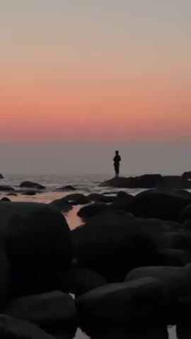 The video shows a silhouette of a person fishing on a rocky outcrop at sunset. The sky is colored with hues of orange and pink. Dark rocks are scattered in the foreground leading towards the ocean, where the person stands with a fishing rod. Small waves can be seen in the background.