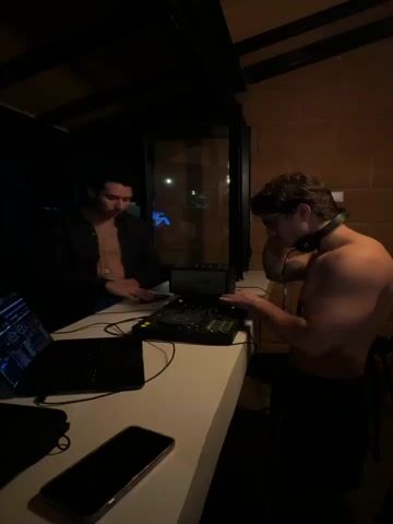 The video captures two men in a dimly lit indoor space, likely a bar or club. One man, standing on the right, is shirtless, wearing headphones, and appears to be operating a DJ mixer. He is focused on the equipment, with his hands on the controls. The other man, standing on the left, is wearing a dark, unbuttoned shirt and holding a bottle. He is looking down at his phone, which is placed on the counter next to a laptop. The background shows a brick wall and a window with some faint lights visible through it. The overall atmosphere suggests a casual, perhaps late-night, DJ session.