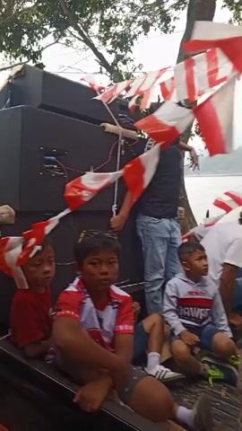 The video shows a street procession with people walking and riding motorcycles. The people are mostly young adults and some children, dressed in casual clothing, some in red and white outfits and some wearing caps. There are flags and banners, some red and white, visible. Large black speakers are mounted on a vehicle, suggesting a musical event or celebration. The procession is moving along a paved walkway next to a body of water, likely a lake or river, with trees and foliage in the background. The sky is overcast, indicating a cloudy day. Some individuals are holding phones, possibly recording the event. The overall atmosphere appears festive and organized.