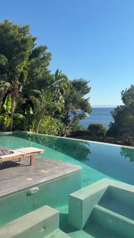 The video shows a clear blue sky and calm ocean waters. In the foreground, there is an infinity pool with turquoise water. A lounge chair with a towel is placed on the pool deck. Lush green trees and foliage surround the pool area. The sun is shining brightly, casting reflections on the water. The scene appears to be a luxurious outdoor setting with a view of the sea.