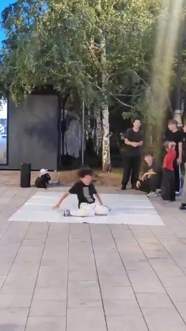 A person in a black shirt and white pants is breakdancing on a white mat in an outdoor plaza. They are performing various moves, including spins and freezes. Several people are watching the performance. The background shows trees and a building.
