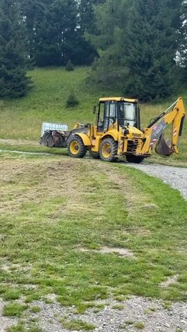 A yellow backhoe loader is parked on a gravel path next to a grassy area. The backhoe has its front loader and backhoe arm raised. The area is a grassy hillside with scattered trees and foliage. The sky appears overcast.