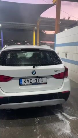 The video shows the rear of a white BMW X1 parked in an outdoor car wash. Water is being sprayed on the car from the left, creating a mist and suds. The car's license plate is visible, reading "KYC 101". The sky in the background is a dusky pink and purple. To the right of the car is a white wall with blue stripes. The ground is wet and soapy. There are no people visible in the frame.