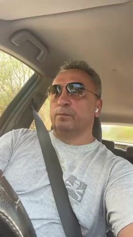 A man is sitting in the driver's seat of a car. He is wearing sunglasses, a grey t-shirt with a skull and crossbones graphic, and a seatbelt. He has short, grey hair and is looking straight ahead. There is a glimpse of trees and sunlight through the car window. He appears to be in motion.