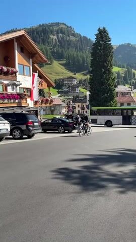 A person on a bicycle rides past a row of parked cars and buildings in a mountain village. The buildings are made of wood with balconies adorned with flowers. The mountains in the background are covered with trees. The sky is clear and blue.