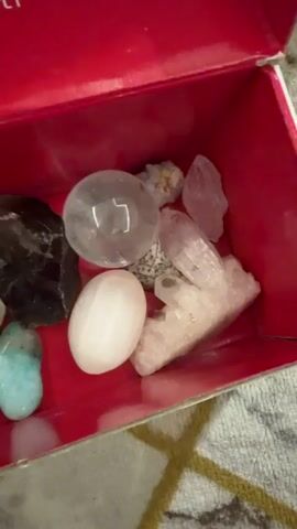 The video begins with a close-up view of a red box containing various objects. The box has white text printed on the inside of its lid, which reads "Love Your Skin, Love Your Soul" and "www.missha.com.tr". Inside the box, there are several stones of different shapes, sizes, and colors, including a large, dark, possibly black stone, a clear sphere, a white oval-shaped object, several light-colored, smooth stones, a turquoise-colored stone, and a pink, smooth object that appears to be a gua sha tool. The camera then pans to the right, showing more of the contents of the box, including some crystalline structures. The video then cuts to a shot of a hand reaching into the box and picking up one of the smaller, white objects. The background behind the box appears to be a patterned fabric, possibly a blanket or rug.