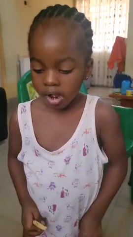 A young Black girl with cornrow braids and small gold earrings, wearing a white tank top patterned with musical instruments, stands indoors. She is facing the camera and slightly looking down, her mouth slightly open. She scratches behind her ear. A green plastic chair is behind her, and in the background is a refrigerator, curtains, and other furniture. The lighting is fairly bright.