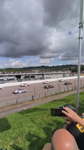 The video shows a race track, with several race cars speeding down the track. The camera is held by a person who is seated on a grassy hill. In the background, a grandstand is visible, filled with spectators. The sky is cloudy, and there is a tall flagpole near the camera.