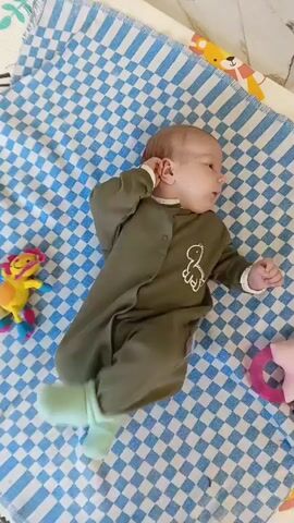 A baby lies on a blue and white checkered blanket. The baby wears a green onesie with a white dinosaur applique. The baby's head is to the left. There are several toys nearby. The baby begins to yawn widely.