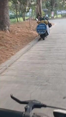 The video begins with a close-up of a bicycle handlebar, indicating the footage is from a first-person perspective, likely on a bike. The camera pans slightly to reveal a paved pathway winding uphill, bordered by a grassy embankment on the left and trees on the right. A motorcycle is parked on the left side of the path, with its kickstand down. Attached to the rear of the motorcycle is a large, circular blue sign with white text. The text on the sign is partially obscured but appears to read "Siena Komet Gas" followed by other characters.

Further up the path, a person is seated on the ground near the base of a large tree, facing away from the camera. They are wearing a hat. A few other motorcycles and a small food cart or stall are visible in the background, situated under a covered structure.

As the camera continues to move forward, the path curves to the right. In the distance, a large banner is mounted on poles. The banner has text on it, including "WELCOME TO MCC STADIUM" and a logo. The overall environment appears to be an outdoor, possibly park-like setting, with paved roads and natural vegetation. The lighting suggests it is daytime, possibly late afternoon due to the soft light.
