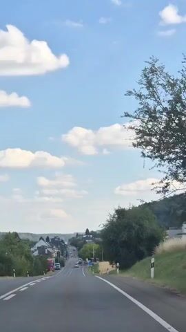 A first-person perspective video shows a car driving on a paved road that curves to the left. The road is bordered by green grass and trees on both sides, with a line of trees and a small town visible in the distance. The sky is blue with scattered white clouds. The sun is shining.