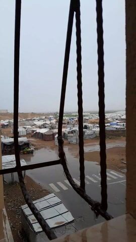 The video, taken from a high vantage point behind a dark metal-barred window, shows a sprawling refugee camp under a gray, overcast sky and rain. The camp consists of numerous makeshift shelters, many covered in white tarpaulin, spread across a flat, brown landscape. Water has pooled on the ground and in the streets, reflecting the gray sky. A paved road with a white crosswalk cuts through the camp in the foreground, and two people are seen crossing the road. The overall scene is bleak and suggests poor living conditions.