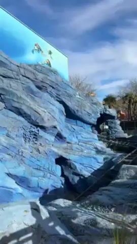 The video shows a roller coaster car ascending a track.  The track appears to be built into an artificial rock formation, which is painted in various shades of blue.  The car is brown and contains several visible people.  Behind the rock formation, palm trees and other theme park structures can be seen.  The sky above is blue with scattered clouds, and a large screen with an image of sea turtles is visible above the rock formation.