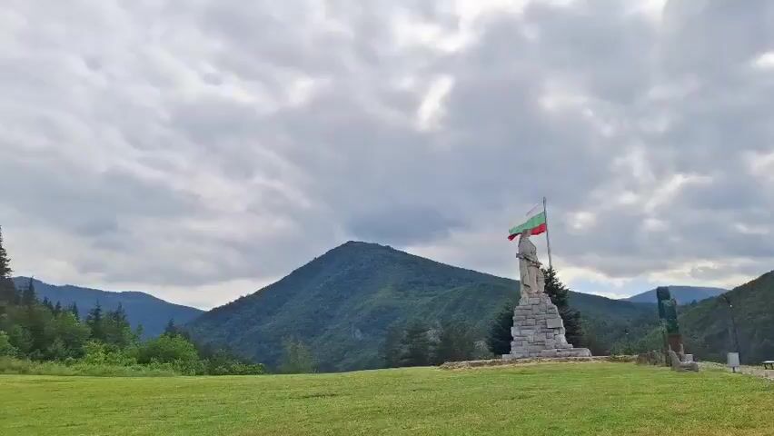 The video opens on a wide shot of a grassy hill with a stone monument topped with a flag. The monument is a rough-hewn stone pillar with a smaller figure on top, and the flag appears to be a tricolor with green, white, and red stripes. Behind the monument, rolling green hills covered in trees form a backdrop, under a sky filled with heavy, grey clouds. The camera then pans slowly to the right, revealing a gravel path with benches and some litter bins along its edge. The path leads towards the edge of the hill, where a valley unfolds. In the distance, a small town with red-tiled roofs is nestled amongst the green hills. The camera continues to pan, showing more of the valley and the surrounding mountainous landscape. The lighting suggests it is daytime, but overcast, with a slightly muted color palette. There are no people visible in the video.