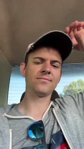 A close-up, head-and-shoulders shot of a young man wearing a baseball cap and a grey hooded sweatshirt. He is in the passenger seat of a car, with the rear window visible behind him. The man is looking at the camera and smiling slightly, with his right hand resting on the back of his head. Sunglasses are clipped to the front of his sweatshirt. The background outside the car appears to be rural, with trees and a clear sky.