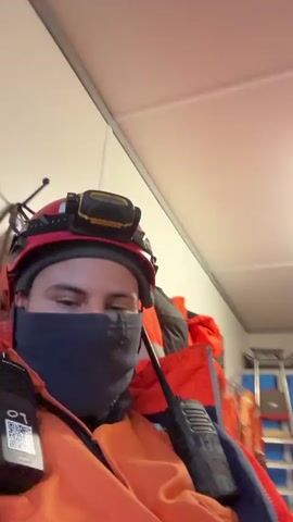 The video shows several people in an indoor room. The people are wearing orange work jumpsuits or orange jackets. The man closest to the camera is wearing a red hardhat with a headlamp attached. He is wearing a black face mask and has two walkie-talkies strapped to his chest. In the background, there are blue lockers, a white board, red chairs, and a table. The room appears to be a break room or meeting room for workers. Some people are sitting while others are standing and talking. One person is looking at a smartphone. The lighting in the room is provided by fluorescent lights.