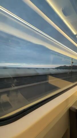 From inside a moving train, the view through the window reveals a landscape rushing by. The window is a multi-pane design with interior reflections of the train's lighting. Outside, a concrete barrier and metal fence separate the train from what appears to be a body of water. Beyond the water, vegetation is visible against a blue sky with clouds. The train is moving quickly, blurring the details of the passing scenery.