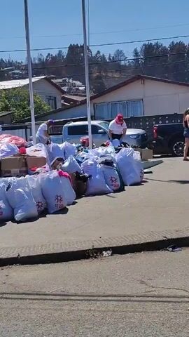 The video shows a sunny outdoor scene with people sorting through piles of donations on a raised platform. The donations appear to consist of clothing, blankets, and other goods, packed in large white bags and cardboard boxes. A white van with "PDI" markings is visible in the background, along with a blue tent. People are seen organizing and moving the donated items. A wooden bench sits near a tree on the platform, where two people are sitting together. The location appears to be a public area, possibly a park or community space. The camera pans across the scene, capturing the extent of the donation effort and the presence of volunteers and support vehicles.