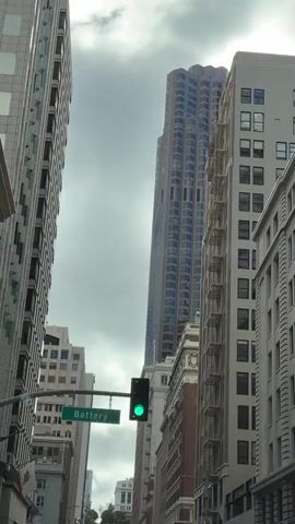 A traffic light cycles from green to yellow then to red as a vehicle travels down a city street lined with tall buildings. A street sign reading "Battery" is visible below the traffic light. The sky is overcast.