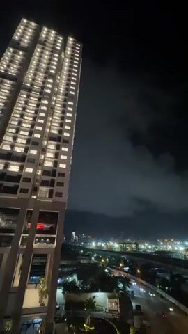 A drone or camera is positioned at a high vantage point, looking down and out over a cityscape at night. In the foreground, on the left, is a portion of a tall, modern, illuminated skyscraper with many windows. Below and in the distance, a city glows with lights. Several roads are visible, with car headlights and taillights indicating movement. A highway overpass or elevated train track crosses the scene. In the far distance, there appear to be port facilities with cranes and numerous lights. Clouds are present in the night sky, obscuring some of the distant lights. The overall impression is of a busy, illuminated urban environment at night.