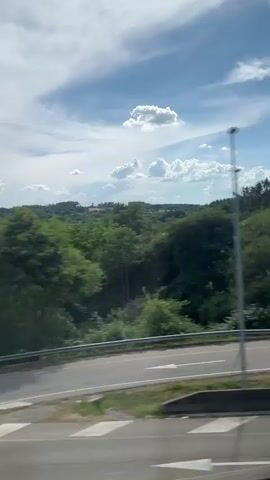 The video shows a view from a moving vehicle looking out at a landscape under a bright blue sky with scattered white clouds. The landscape is predominantly green with rolling hills covered in trees and vegetation. In the foreground, there is a paved road with a white stop line painted on it. Various houses and buildings can be seen nestled within the greenery, some appearing to be older and slightly run-down, others more modern. There are also signs of human presence such as fences, a car, and utility poles. The movement of the vehicle creates a blur effect on the trees and buildings, indicating the perspective of a passenger traveling through the area.