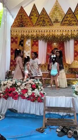 A group of young girls, most under the age of ten, are gathered on a raised stage adorned with elaborate golden and red decorations and a large floral arrangement of red and white flowers. A blue tarp covers the ground in front of the stage. The girls are dressed in a mix of formal and casual attire. One girl wears a light brown formal dress. Several other girls wear light-colored shirts and what appear to be light blue or white undergarments or shorts. One girl in a white shirt with a cartoon print is dancing. Another girl in a light brown dress stands to the side, observing. The overall impression is that of a performance or a celebration.