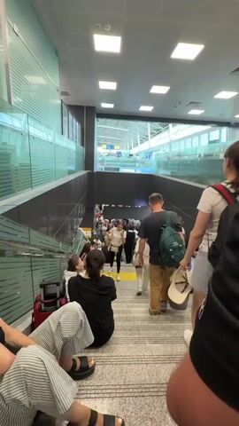 The video shows a busy airport terminal with people waiting in lines. The scene appears to be a descending walkway or staircase leading to a lower level where more people are gathered. Some individuals are sitting on the stairs, others are standing and looking forward. Luggage is visible next to some of the people. The environment is brightly lit with fluorescent ceiling lights. There are glass partitions on the left side of the walkway. In the background, there are signs and store fronts, suggesting commercial areas within the airport. The overall impression is one of a crowded and active travel hub.