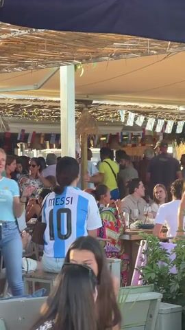 The video shows a crowded outdoor scene, likely a beachside cafe or bar, during the daytime. People are seated at tables and standing around, many wearing casual summer clothing. Several individuals are wearing soccer jerseys, predominantly in the colors of Argentina, with the name "Messi" visible on the back of at least one. The atmosphere appears relaxed and social. There are umbrellas and awnings providing shade. In the background, there's a stage or platform with speakers, suggesting some form of entertainment. Some patrons are interacting with each other, while others are looking at their phones. The ground appears to be a mix of sand and possibly a paved area. Lush green plants are visible in the foreground, adding to the tropical or beachside ambiance. The lighting suggests it's a bright, sunny day.