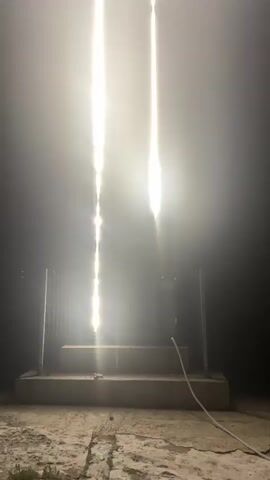 The video shows a large illuminated cross in a foggy environment. The cross is made of white and purple lights. The video starts from ground level looking up at the cross, and the camera pans upwards to reveal the full structure. The cross appears to be made of LED lights. There is a platform or stage in front of the cross, and some fencing on either side. The lighting is very bright, and the fog creates a hazy effect. The overall impression is one of a dramatic and awe-inspiring display.