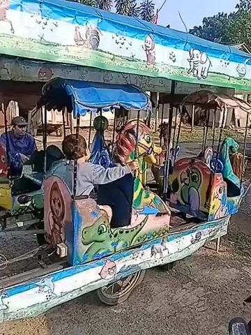 A small, colorful amusement ride is shown, likely a carousel or a train-like ride for children. The ride is decorated with various cartoon characters and animals, including dinosaurs, elephants, horses, and what appear to be simplified human figures. A young child is seated in one of the ride's compartments, holding onto a control or decoration. The ride appears to be outdoors, with a dirt or gravel ground visible. The background suggests a rural or semi-rural setting, with some structures and trees. The ride itself is mounted on wheels, implying it is mobile. The overall impression is of a simple, rustic amusement intended for young children.