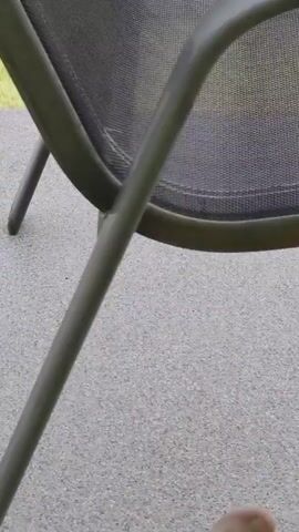 The video shows a close-up of a black mesh outdoor chair with a metal frame. The camera pans to the right, revealing a metal railing, green grass, and a portion of a building with an orange facade and a tiled roof. Further in the background, a large, rocky mountain covered in trees dominates the landscape under a cloudy sky. The scene appears to be a view from a balcony or patio.
