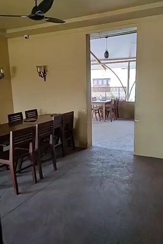 The video shows a building's interior transitioning to an outdoor terrace. Inside, a dining area is visible with a long wooden table surrounded by chairs, under a ceiling fan and wall-mounted light fixtures. The wall is a light yellowish-brown. The floor is concrete. Beyond a large opening, an outdoor terrace can be seen. The terrace contains matching wooden tables and chairs arranged to accommodate a large group. Bottles of water and plates are set on the tables. The terrace is covered with a roof supported by pillars, and beyond it, the outside cityscape is visible. The lighting shifts from dim inside to brighter on the terrace.